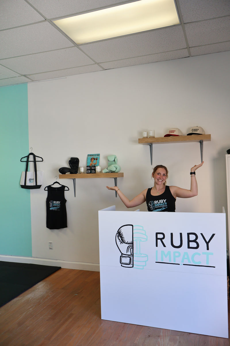 A woman stands behind a white reception desk in a fitness studio, wearing a black tank top with the 'Ruby Impact' logo. The room features a white and light teal wall, wooden floating shelves with various items, and a wood floor.