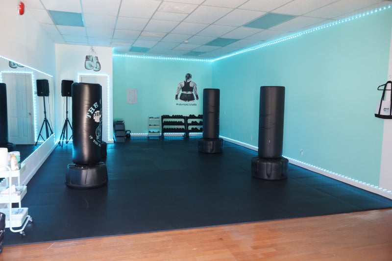 A gym or training room with three black cylindrical punching bags on a black mat, light blue walls, LED strip lighting, and a wooden floor.