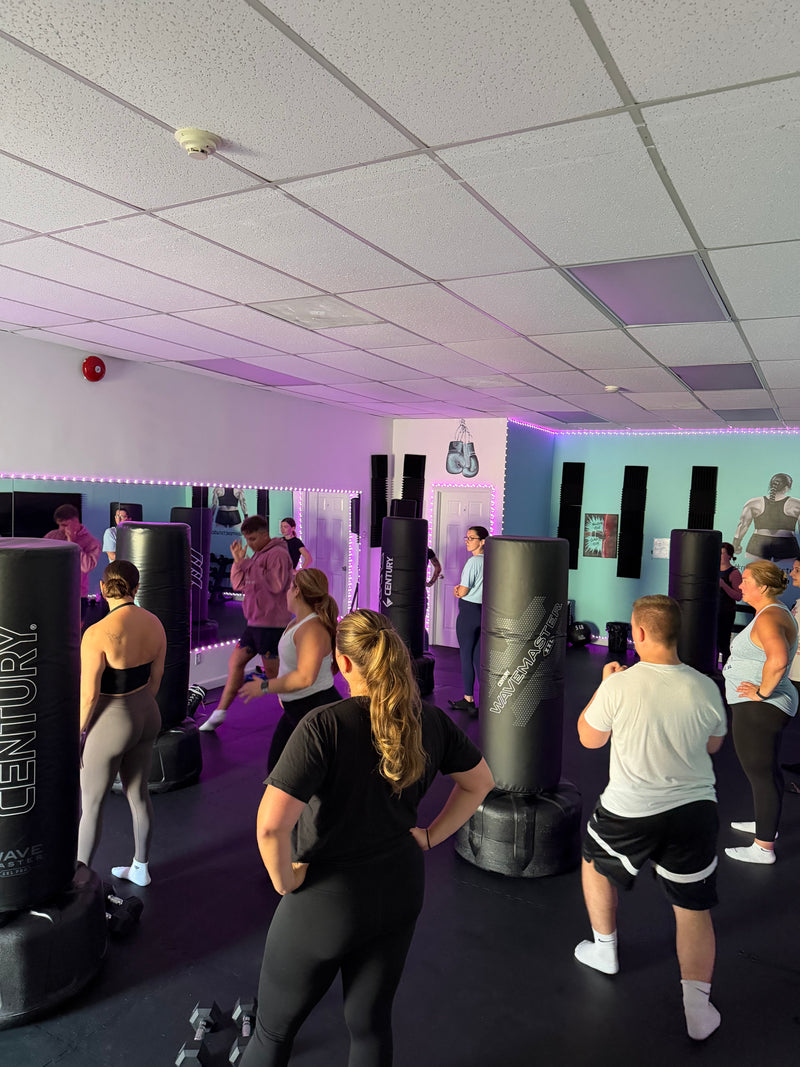 A group of people training in a gym with multiple black punching bags, including a prominent Century brand bag in the foreground, under purple and pink ambient lighting.