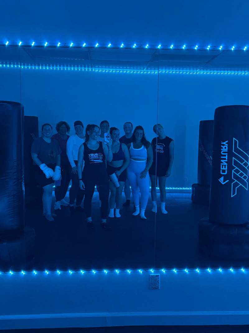 A group of people standing together in a gym or training space with blue LED lighting, with punching bags visible on both sides.